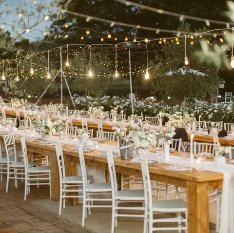 Long festoon-lit reception table at dusk, dressed with ivory florals and olive foliage