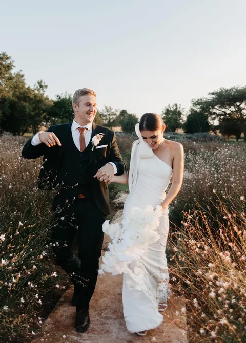 Couple walking through a dried-grass meadow with a white rose bouquet